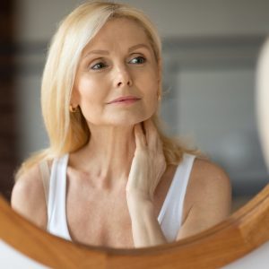 Beautiful aged european woman touching neck and looking at mirror, making anti-aging beauty routine