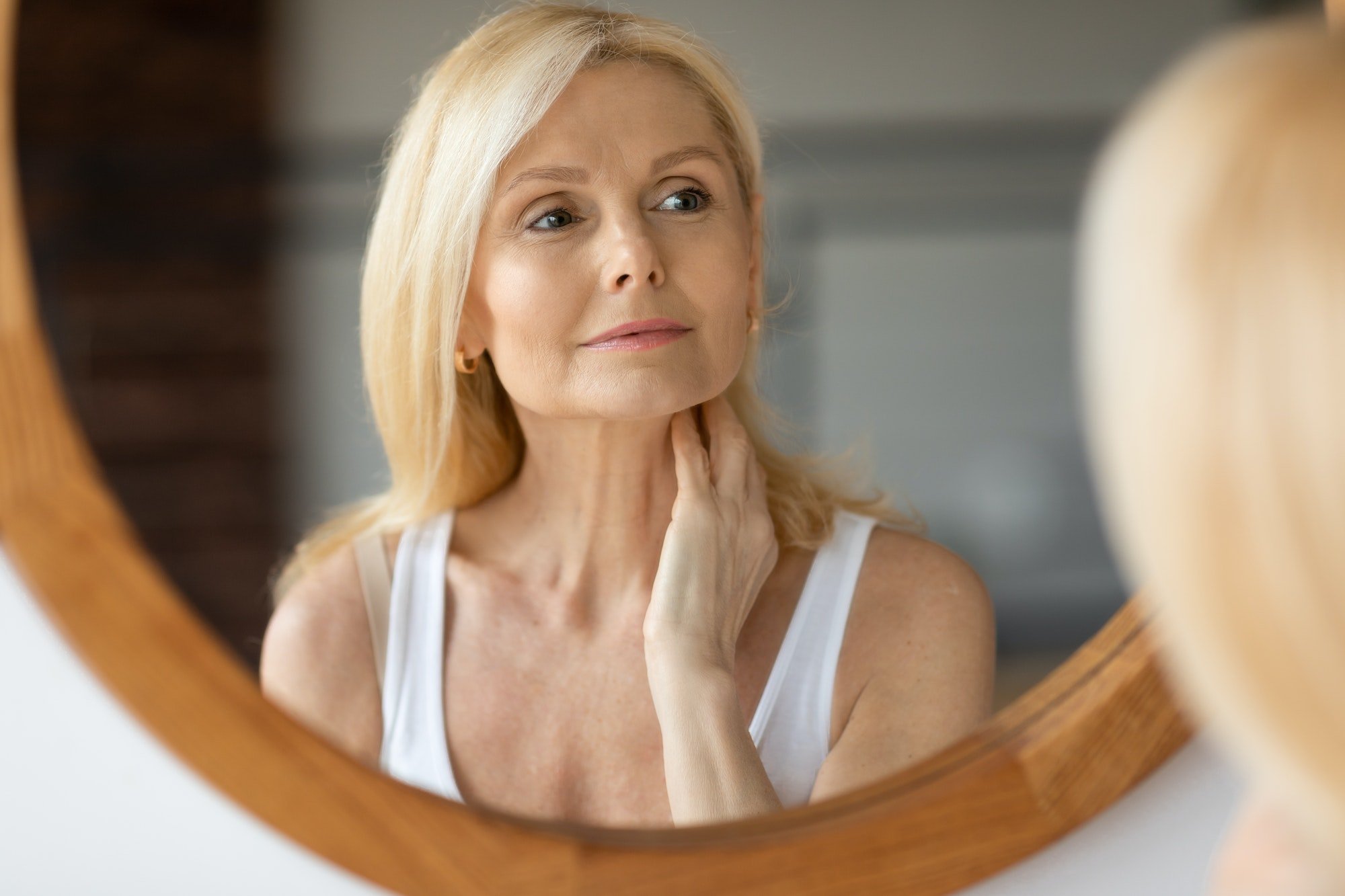 Beautiful aged european woman touching neck and looking at mirror, making anti-aging beauty routine