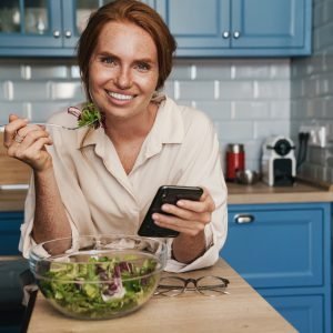 Image of smiling redhead woman using cellphone while eating salad
