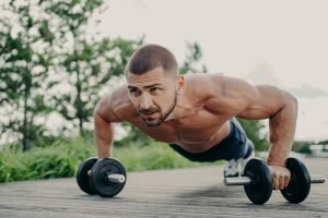 Young motivated sportsman stands in plank pose, trains muscles with barbells