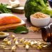 Front view of many fish oil capsules spilling out from the bottle surrounded by an assortment of food rich in omega-3 such as salmon, flax seeds, broccoli, sardines, spinach, olives and olive oil.