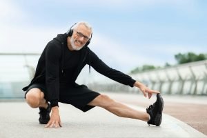 Cheerful healthy elderly man stretching legs in park with music