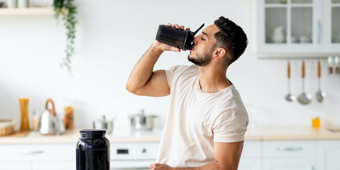 Young Arab guy drinking protein shake from bottle at kitchen, copy space. Body care concept