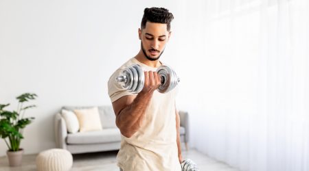 Domestic training concept. Strong young Arab man exercising with dumbbells at home