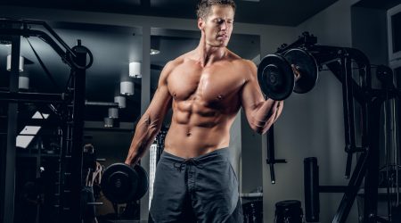 Muscular shirtless male doing biceps exercise with dumbbells.